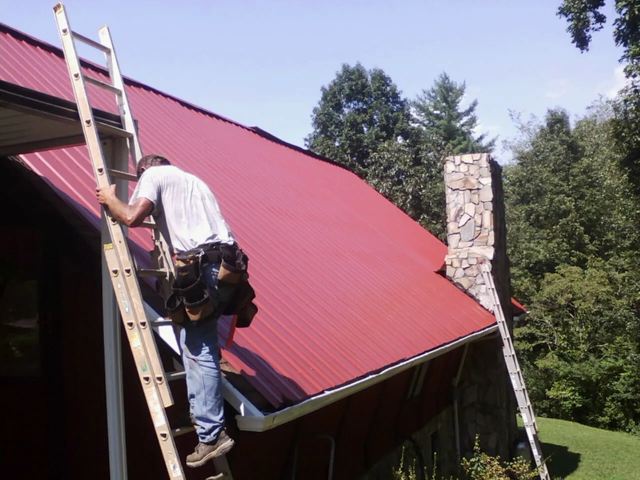 Roofer on a ladder beside a red metal roof and stone chimney — Soffit & Fascia Repair by Roofing Brothers in Mackey